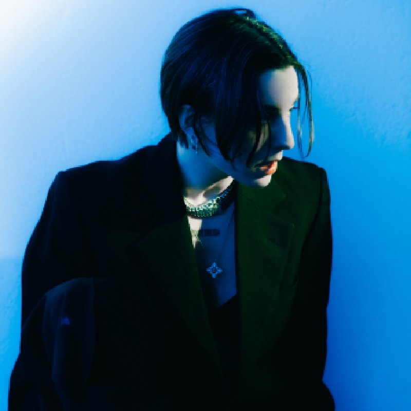 A woman with short hair wearing a suit jacket and silver jewellery looking to the right against a blue background