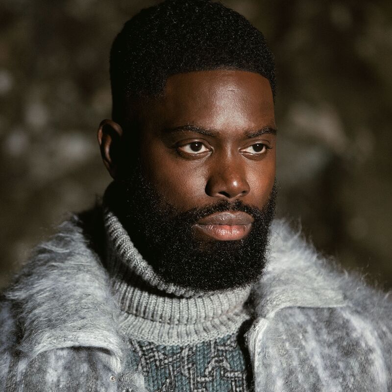 Ghetts wears a light blue coat against a dark background.