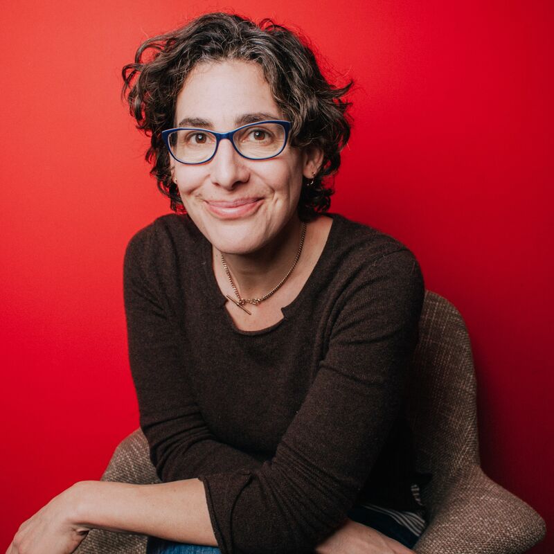 Sarah Koenig sits in front of a red background wearing a grey top