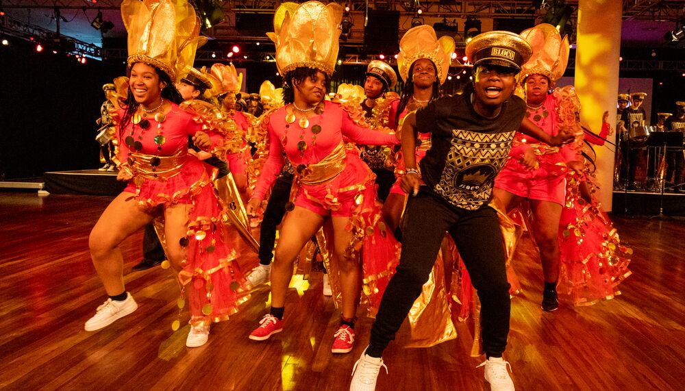 A group of exuberant young dancers from Kinetika Bloco joyfully dance in colorful red and gold carnival outfits.