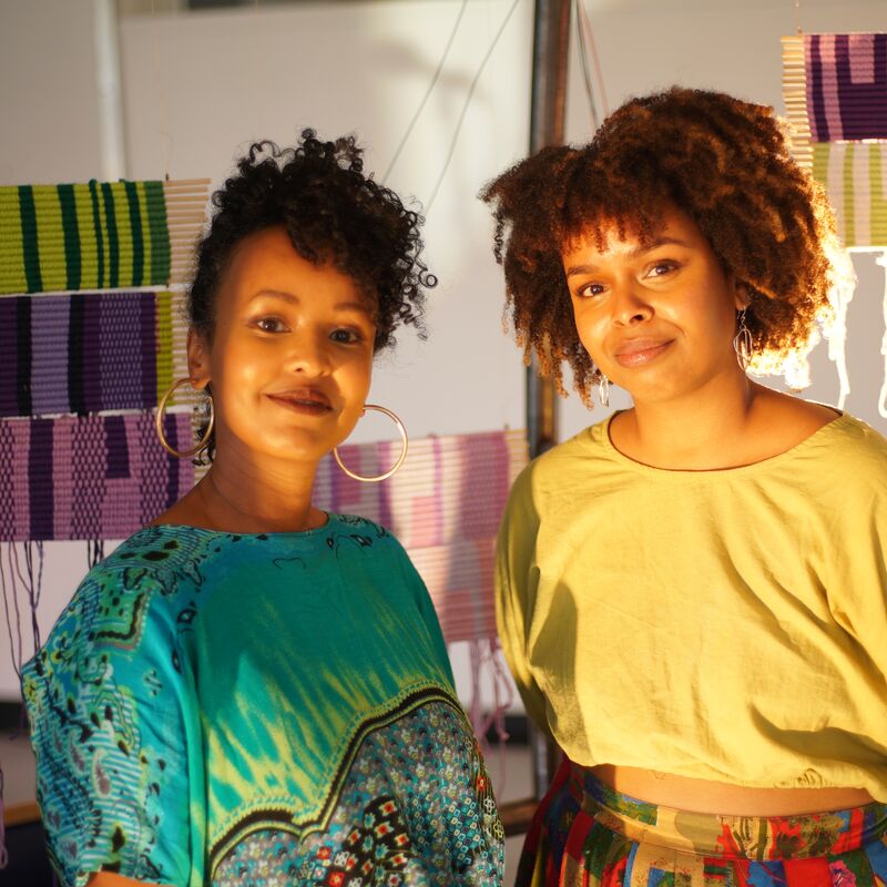 Two women stood in front of pieces of Somalli weaving art