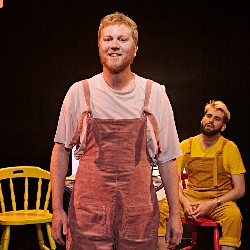 Person wearing pink dungarees stands up looking outwards and smiling. A person wearing yellow dungarees sits behind them.