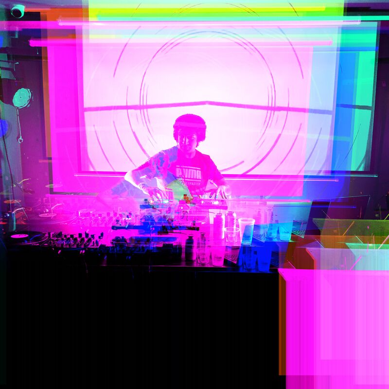 A DJ stands at decks with blue, green and pink lights bouncing round the image.