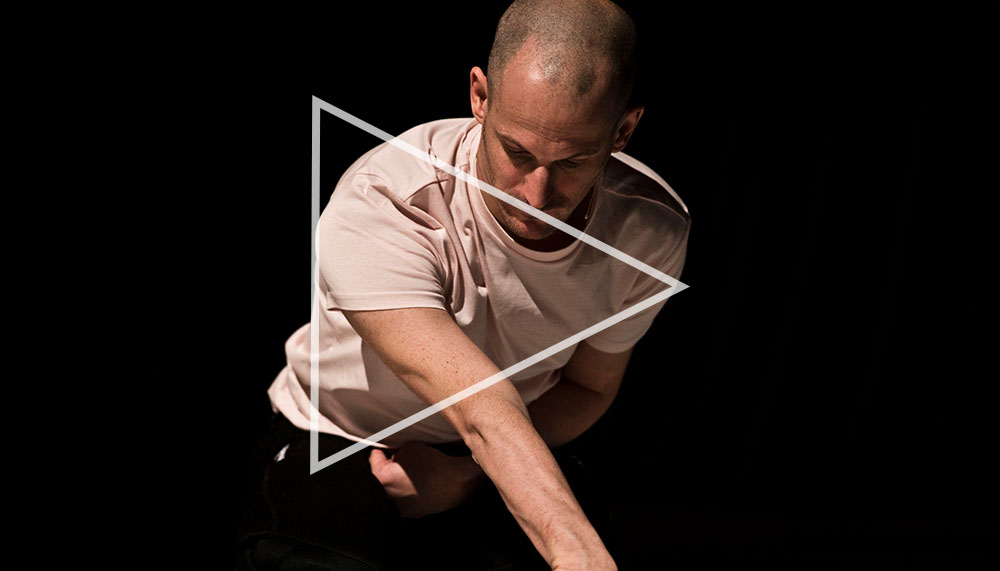 Michael Turinsky kneels on the floor with their arm outstretched in front of them, their wrist bent so that their hand rests on the floor in front of a green toy. They wear a white T-shirt and black trousers.