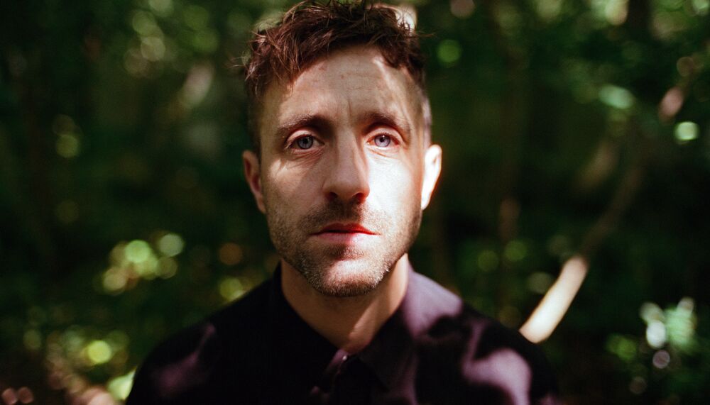 The performance artist Martin O'Brien, a middled aged White man with brown hair shaven at the sides wearing a dark shirt. Martin faces the camera, his face partly obscured by the shadows of surrounding trees