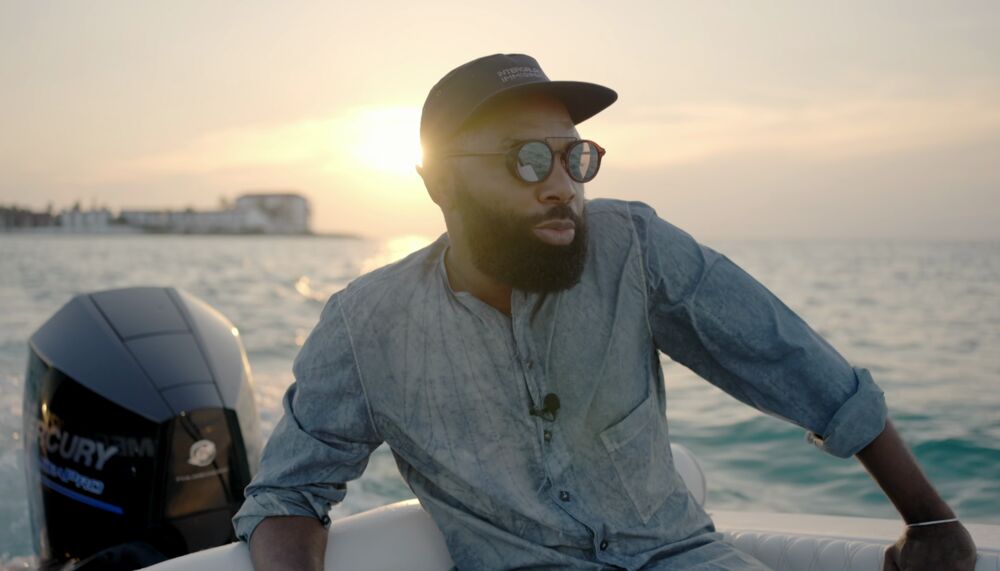 Arist Tavares Strachan, a Black man in his thirties with a thick but well kept beard, wearing a baseball cap and sunglasses, he's sitting at the back of a speed boat, the sun is setting over the sea behind him