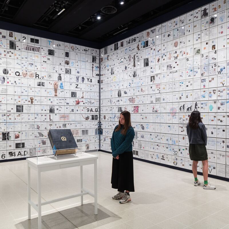 Installation view of a large encyclopaedia, in a room with walls covered in paper clippings.