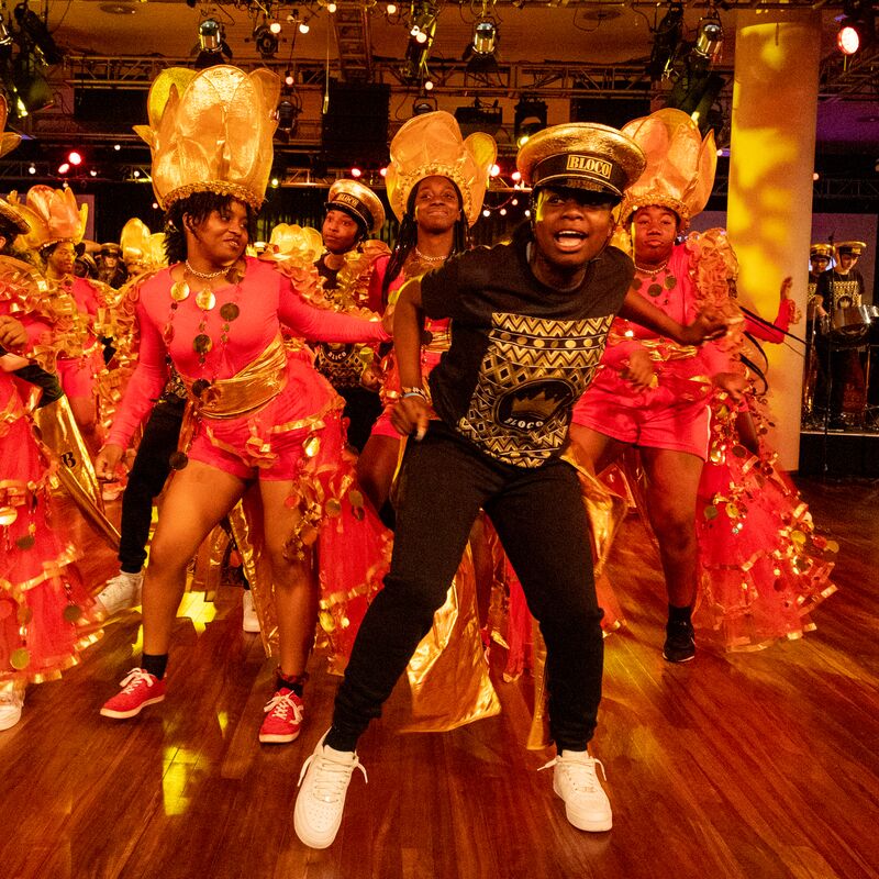 A group of exuberant young dancers from Kinetika Bloco joyfully dance in colorful red and gold carnival outfits.