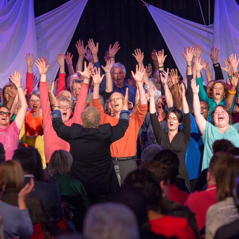 A choir with their arms up in the air, wearing bright coloured t shirts