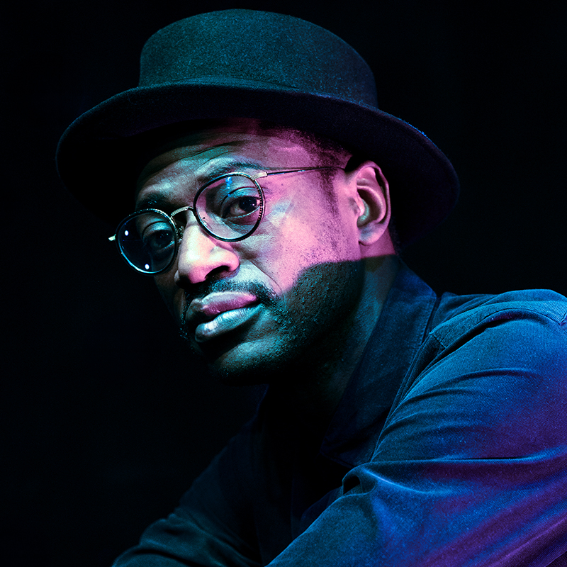 Artist Mark Kavuma wears a hat and black rimmed glasses as he poses for a photo in a dark room