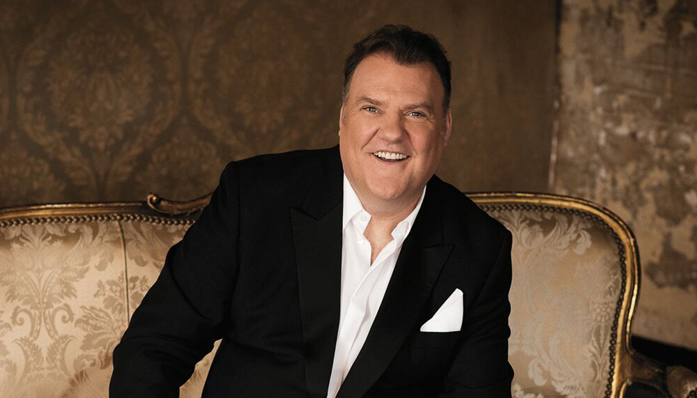 Welsh opera singer Bryn Terfel smiles into the camera dressed in a suit. He is sat on an ornate antique loveseat surrounded by sheets of music.