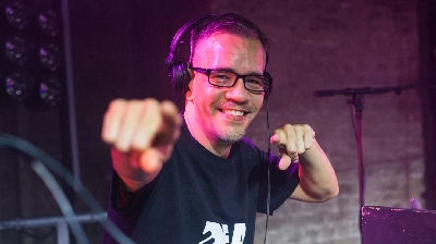 DJ Chinaman stands at decks with headphones on and points and smiles to the camera.