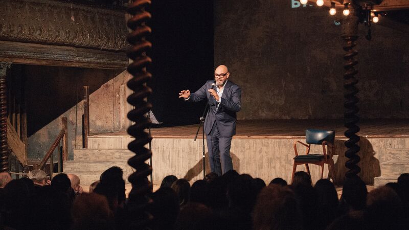 A performer wearing a suit stands on a wooden stage speaking into a microphone to a large audience