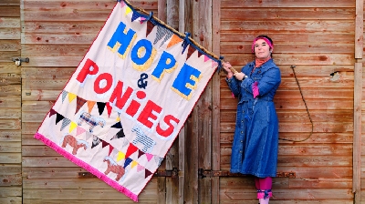 Katherina, a white woman in a denim dress holds a colourful flag that reads "Hope & Ponies" with images of bunting and ponies on.