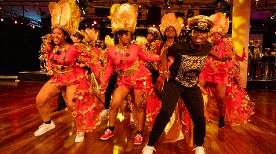 A group of exuberant young dancers from Kinetika Bloco joyfully dance in colorful red and gold carnival outfits.