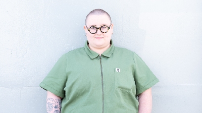 Maddie Morris stands against a white background wearing a green short sleeved jumpsuit and round glasses. They have pale skin, tattoos on one arm, and a shaved head.