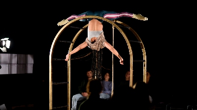 A performer hands upside down while holding on with their legs on a gold arched frame.