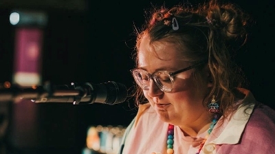 A person wearing a colourful jumper sits at a keyboard with a microphone playing.