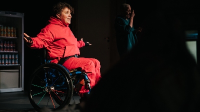 Person wearing a red tracksuit sits in a wheelchair on stage with their arms outstretched beside them.