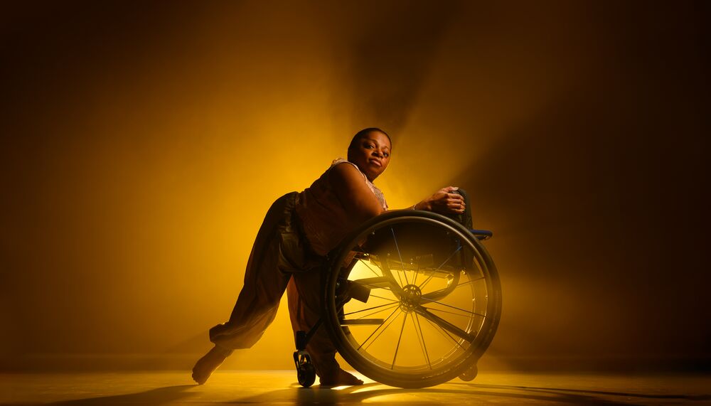 A black female dancer is leaning against her wheelchair facing towards it, in front of an amber light