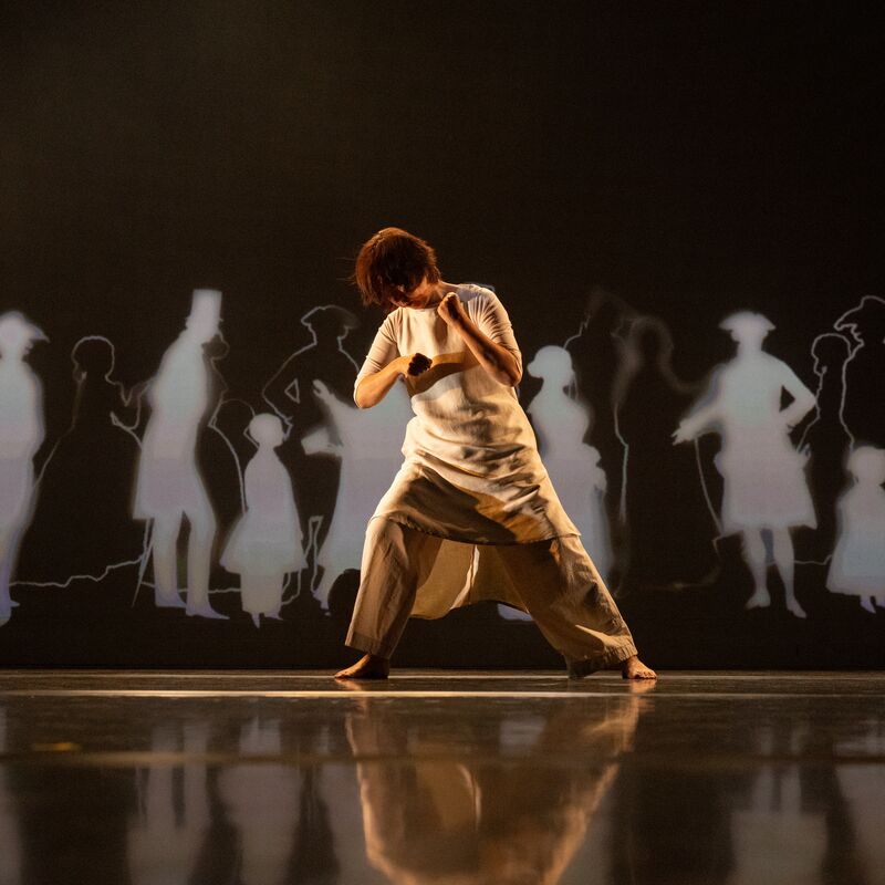 Chisato Minamimura dancing on stage wearing a loose-fitting cream tunic and trousers. Black and white silhouettes of figures in historical costume are projected on the wall behind her.