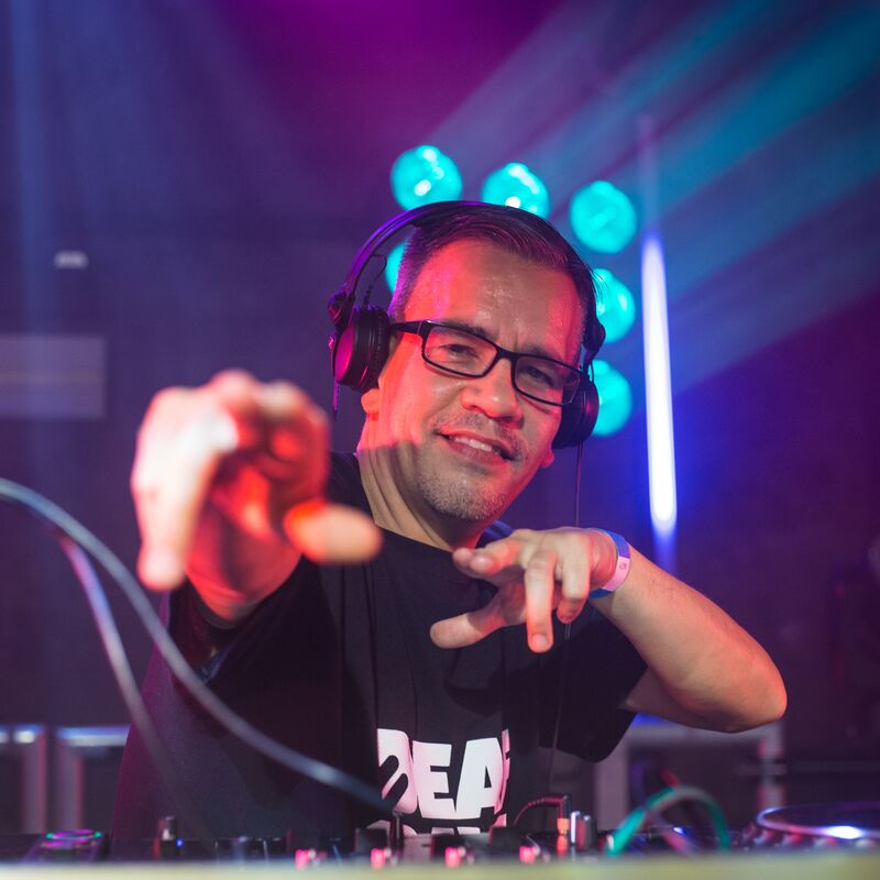 DJ Chinaman stands at decks with headphones on pointing at the camera.