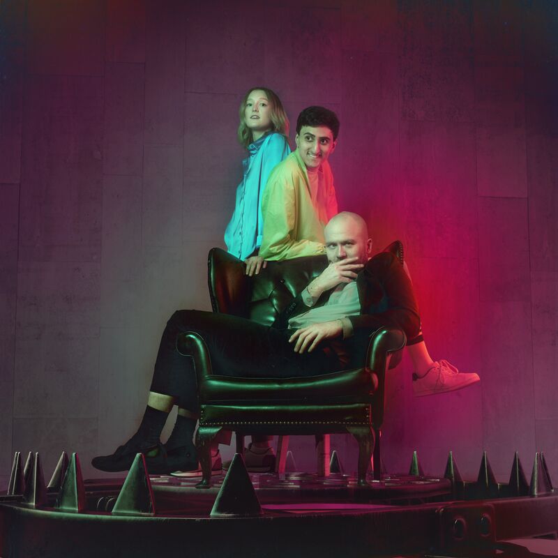 The three people are gathered around a large armchair in an open bear trap against a concrete background illuminated in purple and red.