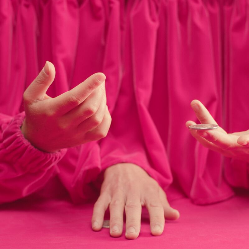 A close up, landscape shot of three white hands emerging from underneath a pink, velvet curtain. The frame is taken up by the pink of the curtain, which is the same material lining the table upon which one of the three hands rests..