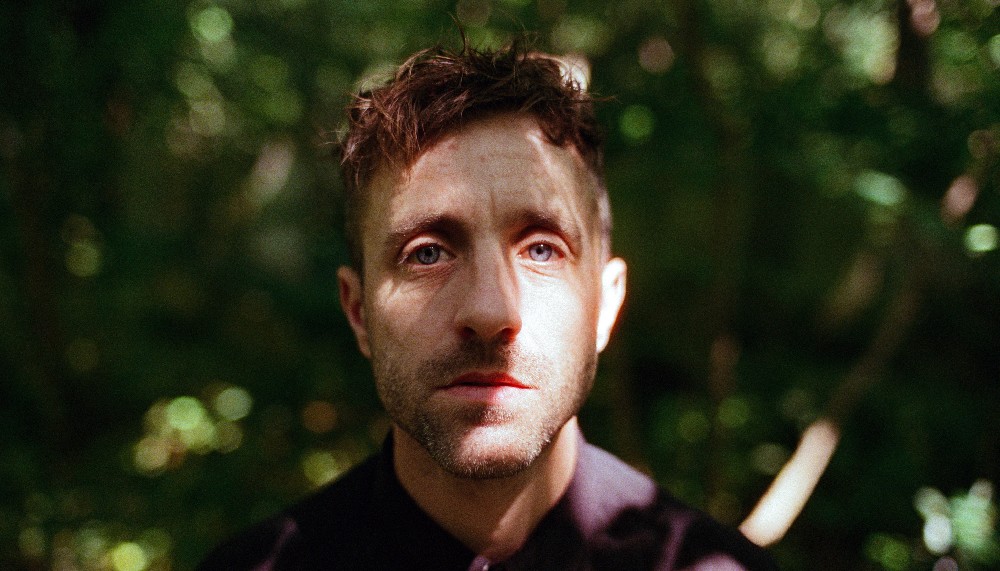 The performance artist Martin O'Brien, a middled aged White man with brown hair shaven at the sides wearing a dark shirt. Martin faces the camera, his face partly obscured by the shadows of surrounding trees.