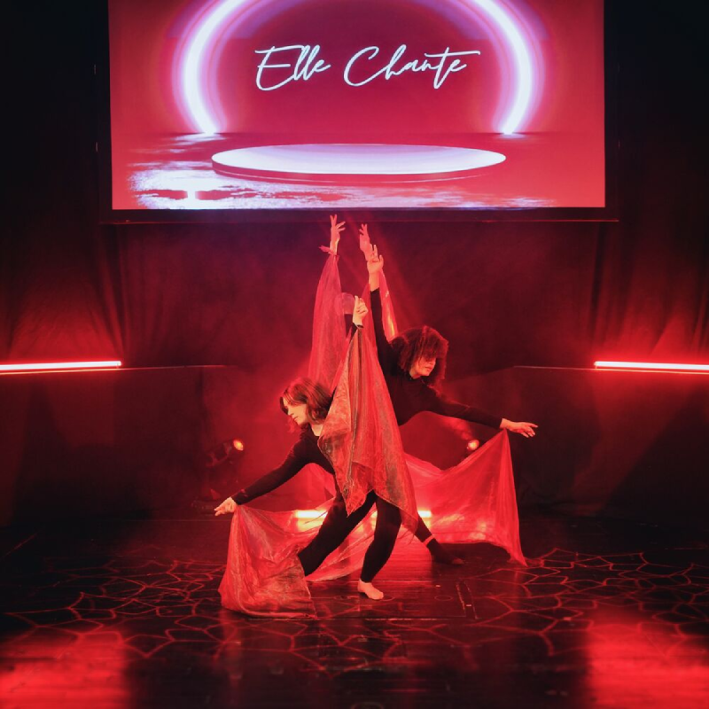 Two dancers wearing all black bend over with their arms outstretched. They have silk scarfs tied to their wrists, and the fabric hangs down to the stage. A screen is behind them with 'Elle Chante' projected onto it. The lighting is red.