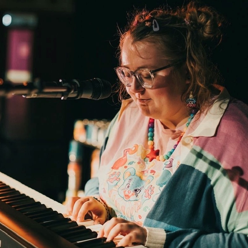 A person wearing a colourful jumper sits at a keyboard with a microphone playing.