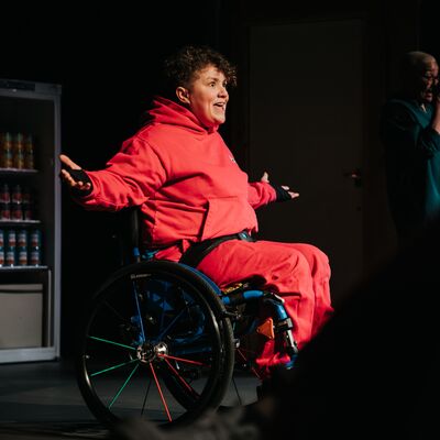 Person wearing a red tracksuit sits in a wheelchair on stage with their arms outstretched beside them.