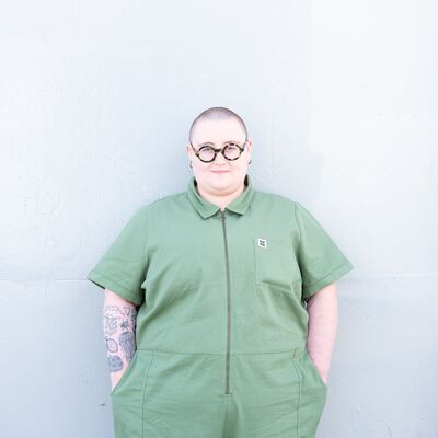 Maddie Morris stands against a white background wearing a green short sleeved jumpsuit and round glasses. They have pale skin, tattoos on one arm, and a shaved head.