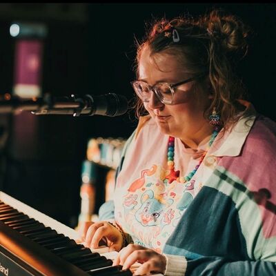 A person wearing a colourful jumper sits at a keyboard with a microphone playing.