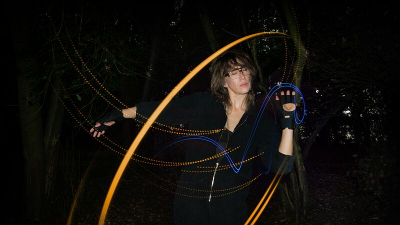 Someone wearing all black with long hair and glasses draws with blue and yellow light in front of a dark background