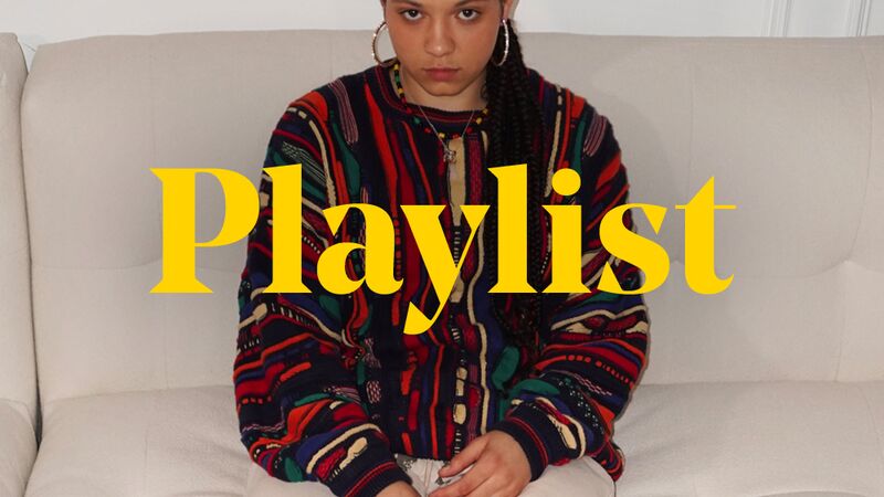 Jaya who wears a patterned top, white trousers and boots is sitting on a white sofa with legs outstretched. The word ‘Playlist’ is emblazoned over the top of the image in yellow type.