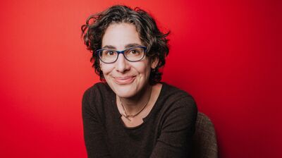 Sarah Koenig sits in front of a red background wearing a grey top