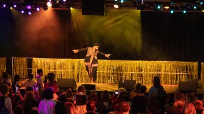 Performer on stage in colourful clothing with their arms straight out to the side