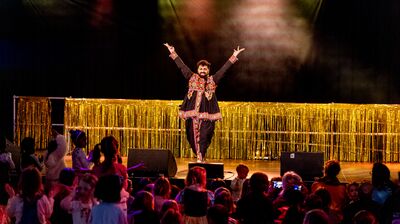 Performer in colorful attire on stage with arms raised, entertaining a crowd of children at an event