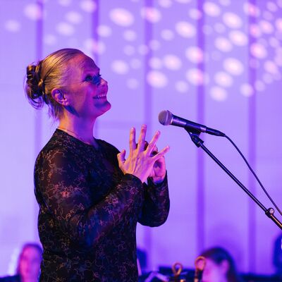 Sigrún Sævarsdóttir-Griffiths smiling, facing a microphone, with a purple background, wearing a black dress.