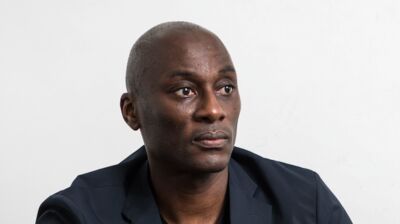 Writer and Curator Ekow Eshun wears a blue suit jacket and sits in front of a white background.