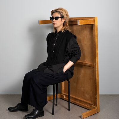Haegue Yang in a black outfit sat on a stool, with a wooden table balancing behind her
