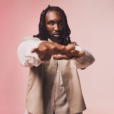 A man with braided hair posing with both hands outstretched wearing white and beige layered shirts