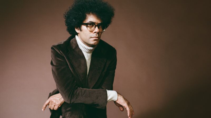 Richard Ayoade wearing a beige turtleneck, brown jacket and glasses