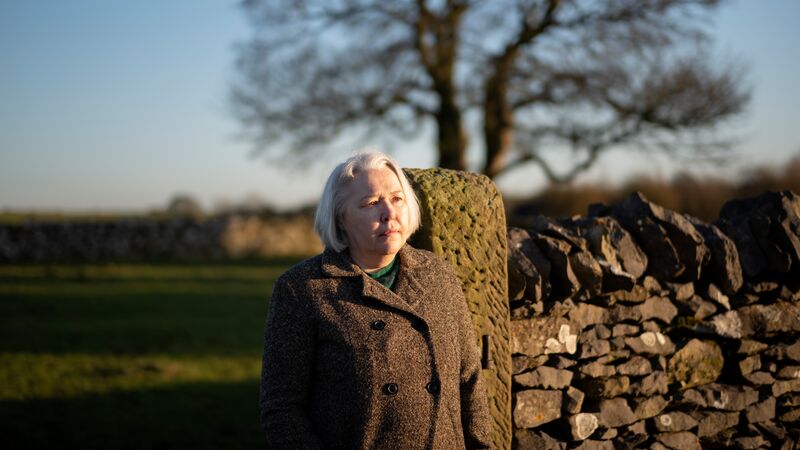 Susanna Clarke in a brown jacket stands by a wall in a field.