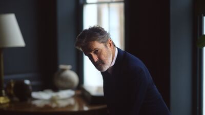 Rupert Everett sits in a dark room, leaning forward and wearing a navy blazer.