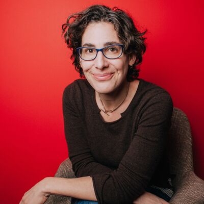 Sarah Koenig sits in front of a red background wearing a grey top