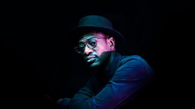 Artist Mark Kavuma wears a hat and black rimmed glasses as he poses for a photo in a dark room