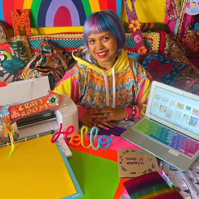 A person sits in brightly coloured hoodie with blue and purple hair surrounded by bright and colourful craft materials