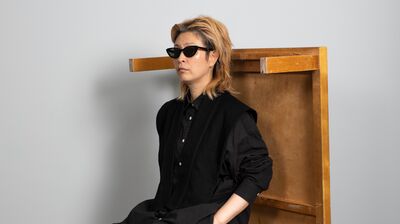 Haegue Yang in a black outfit sat on a stool, with a wooden table balancing behind her